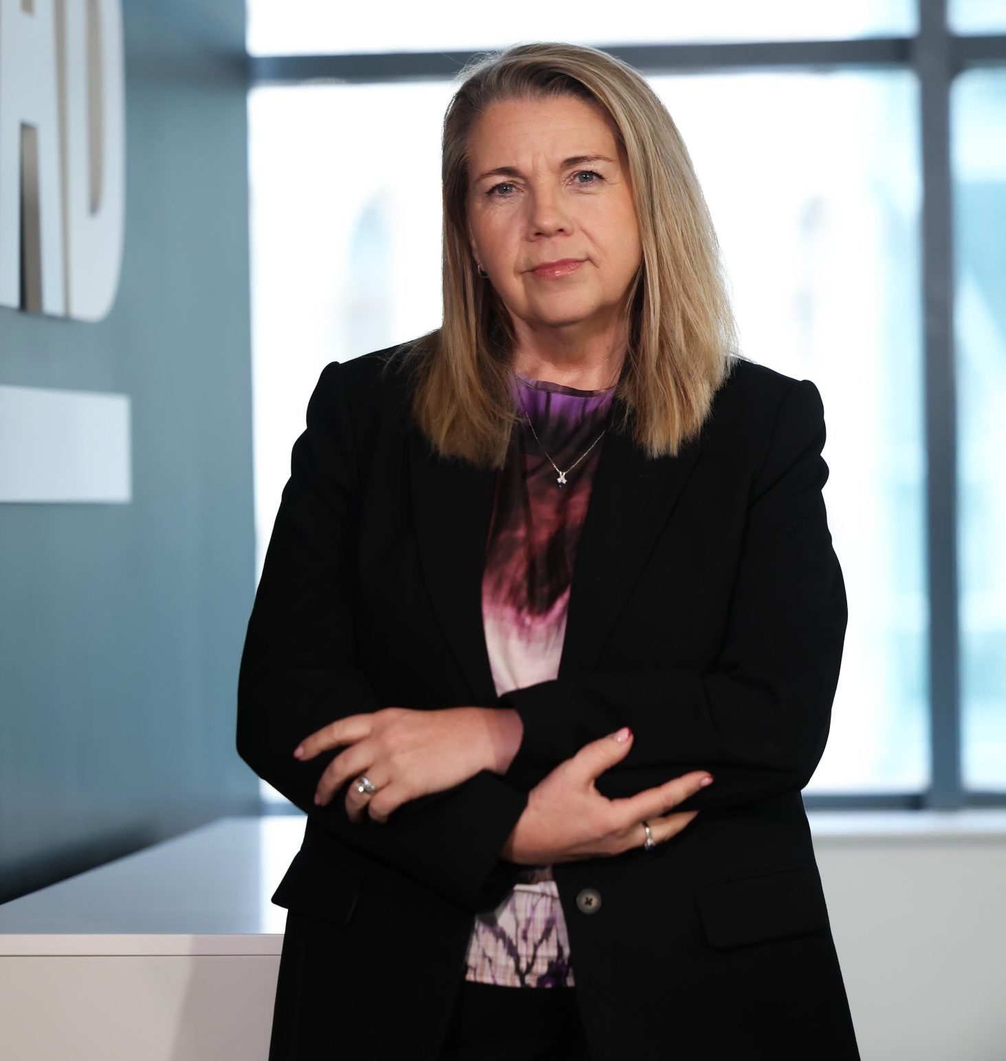 An image of Michelle Hawkins standing in the office wearing a black blazer and purple top. She has her arms folded.