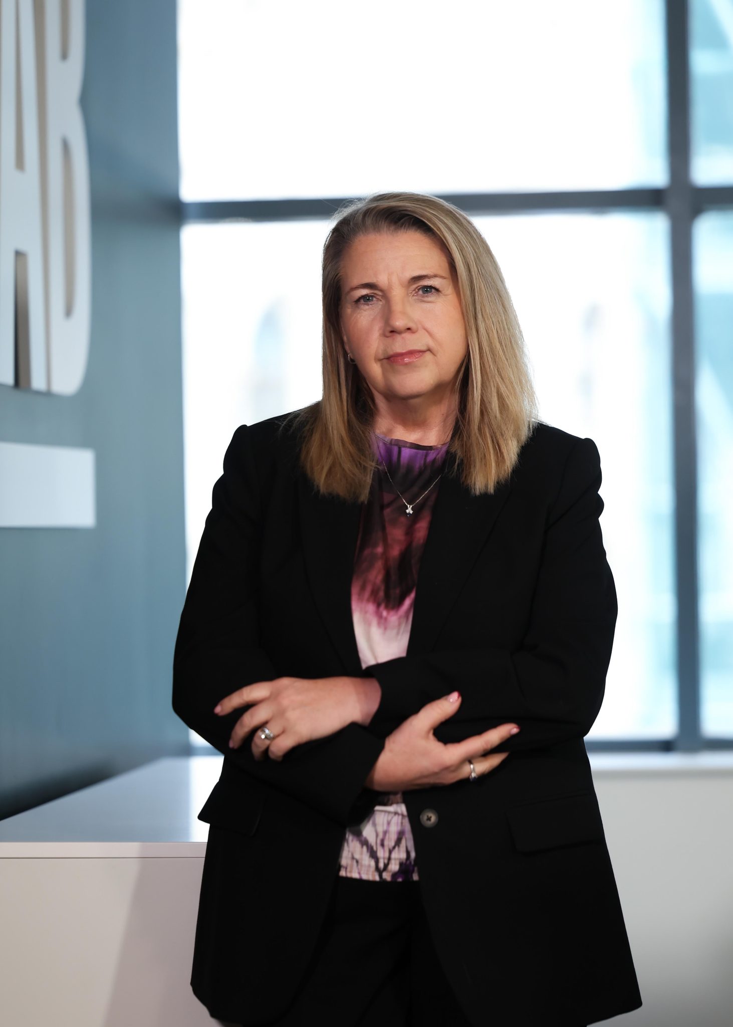 An image of Michelle Hawkins standing in the office wearing a black blazer and purple top. She has her arms folded.