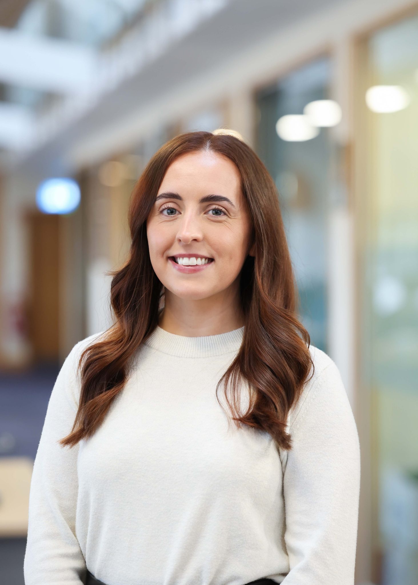 An image of Lauren McMahon wearing a white top. She is standing in the office and smiling at the camera.