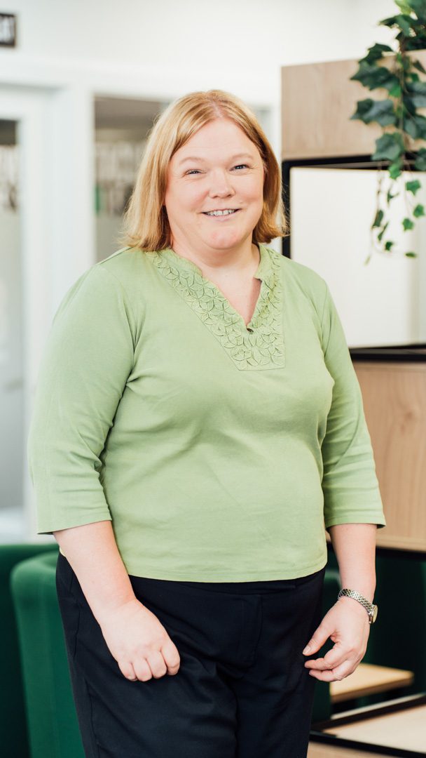 An image of Susan McKechnie standing in the office wearing black trousers and a green top.