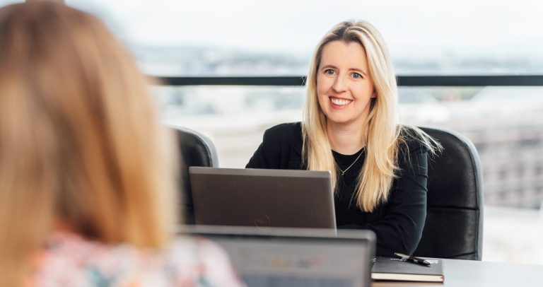 AAB team member on smiling at colleague across meeting room next to laptops.