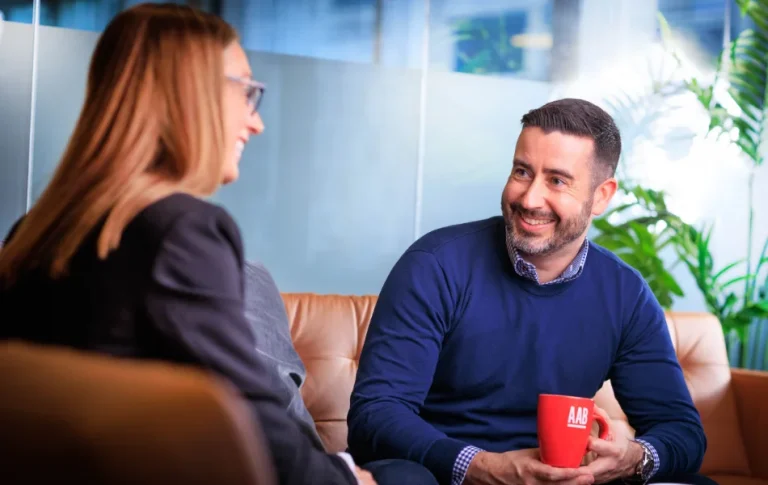 AAB team members David Purse and Jess Constable sitting chatting on couch with red mugs.