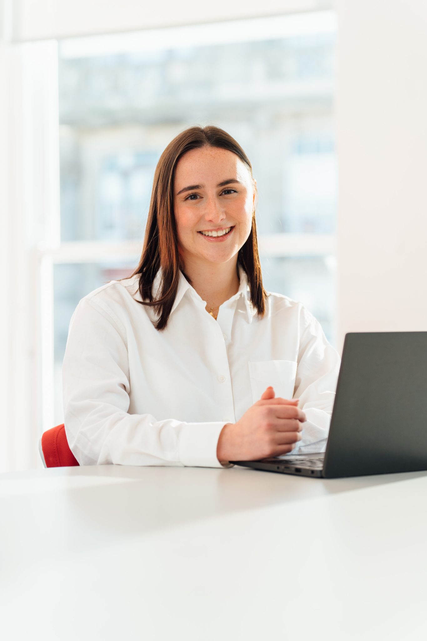 An image of a person smiling at the camera whilst sitting on a desk with her laptop.