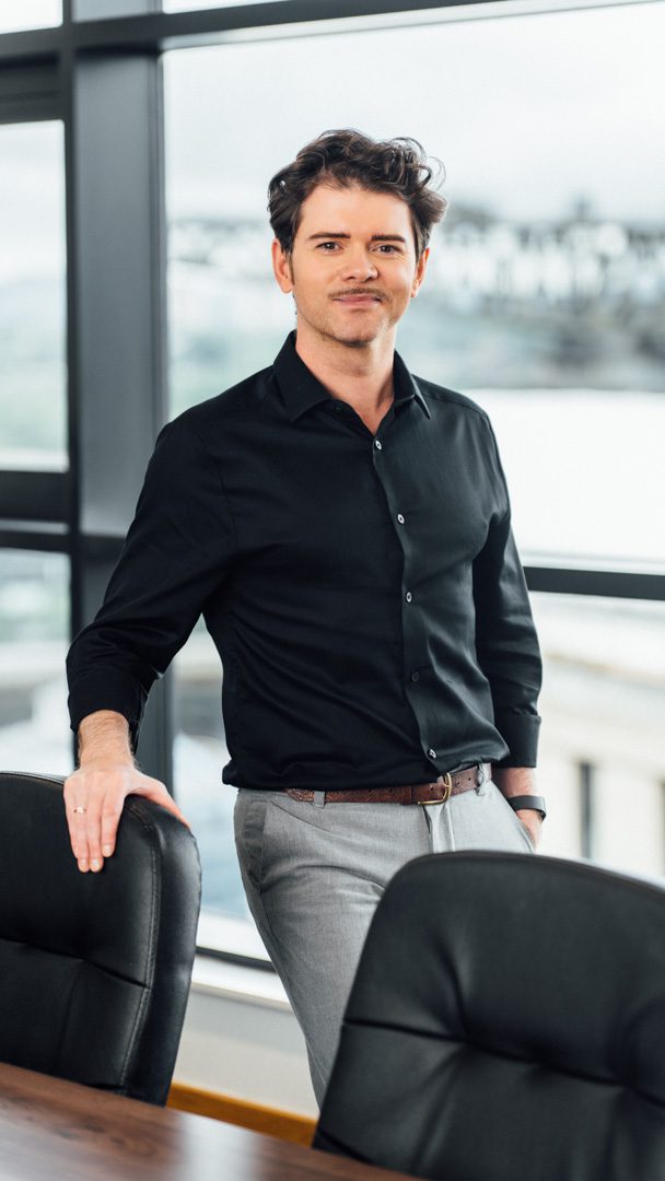 An image of Shaun Harper in the Edinburgh office wearing a black shirt and grey trousers. He is standing next to the window and holding onto a chair.