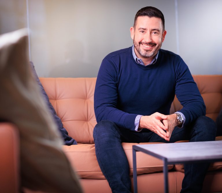 An image of an AAB employee wearing a navy jumper and sitting on a pink couch.