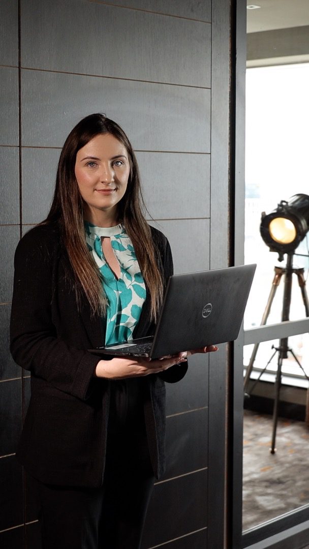 Emma Maginn standing in office holding laptop looking at camera
