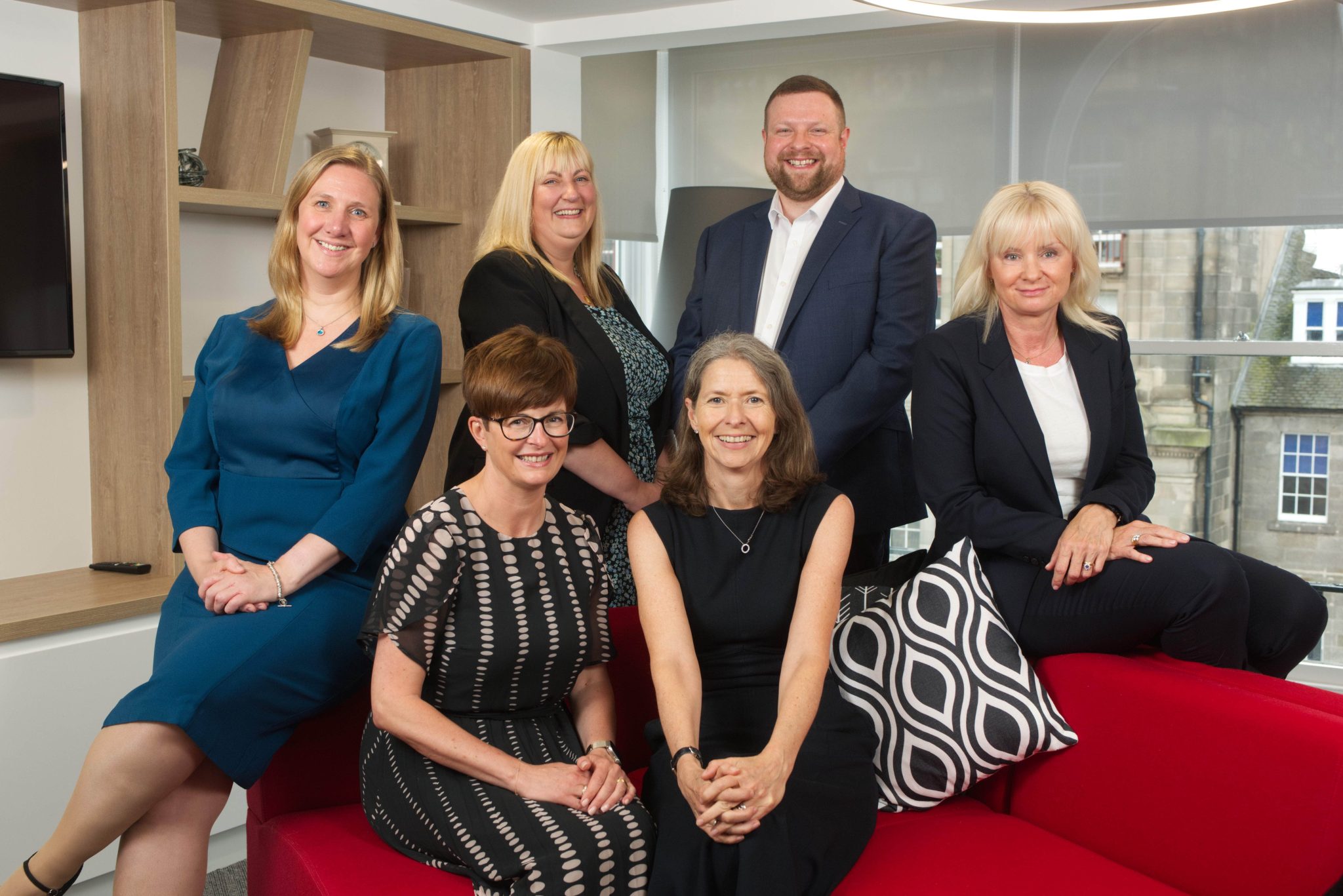 A n image of Emma Lancaster with five other AAB team members who have all been promoted within their roles. They are sitting on a red couch in the office.