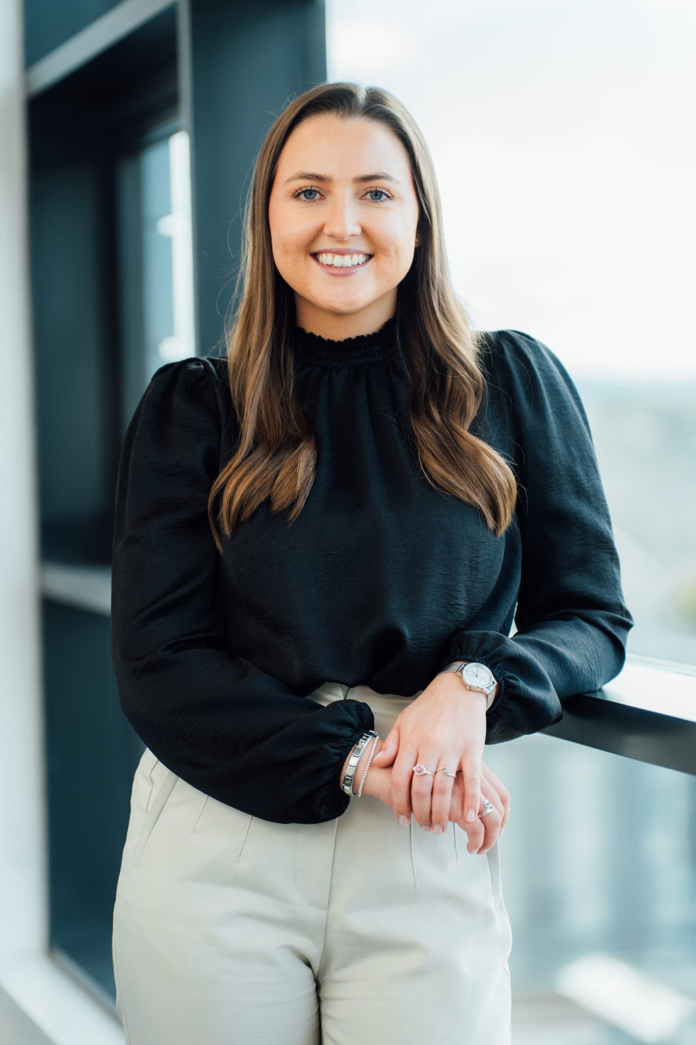 An image of Lori Young standing against a windowsill wearing a black top and cream trousers.