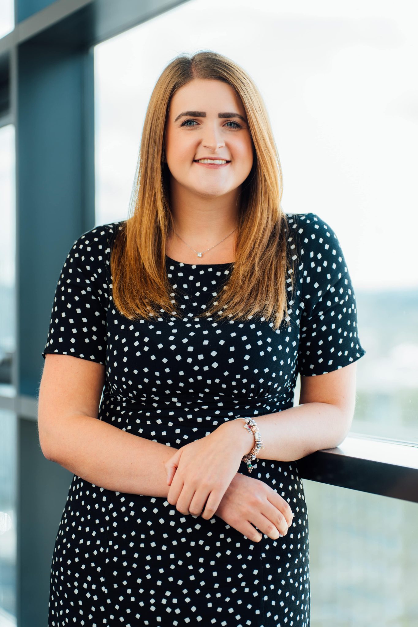 An image of Stephanie Lalley wearing a polka dot dress and smiling at the camera whilst leaning against a window.