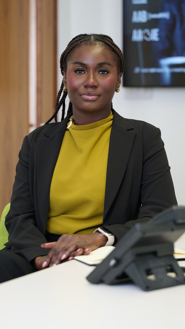 An image of Rosemary Addo wearing a yellow top with a brown blazer. She is sitting at her desk smiling at the camera.