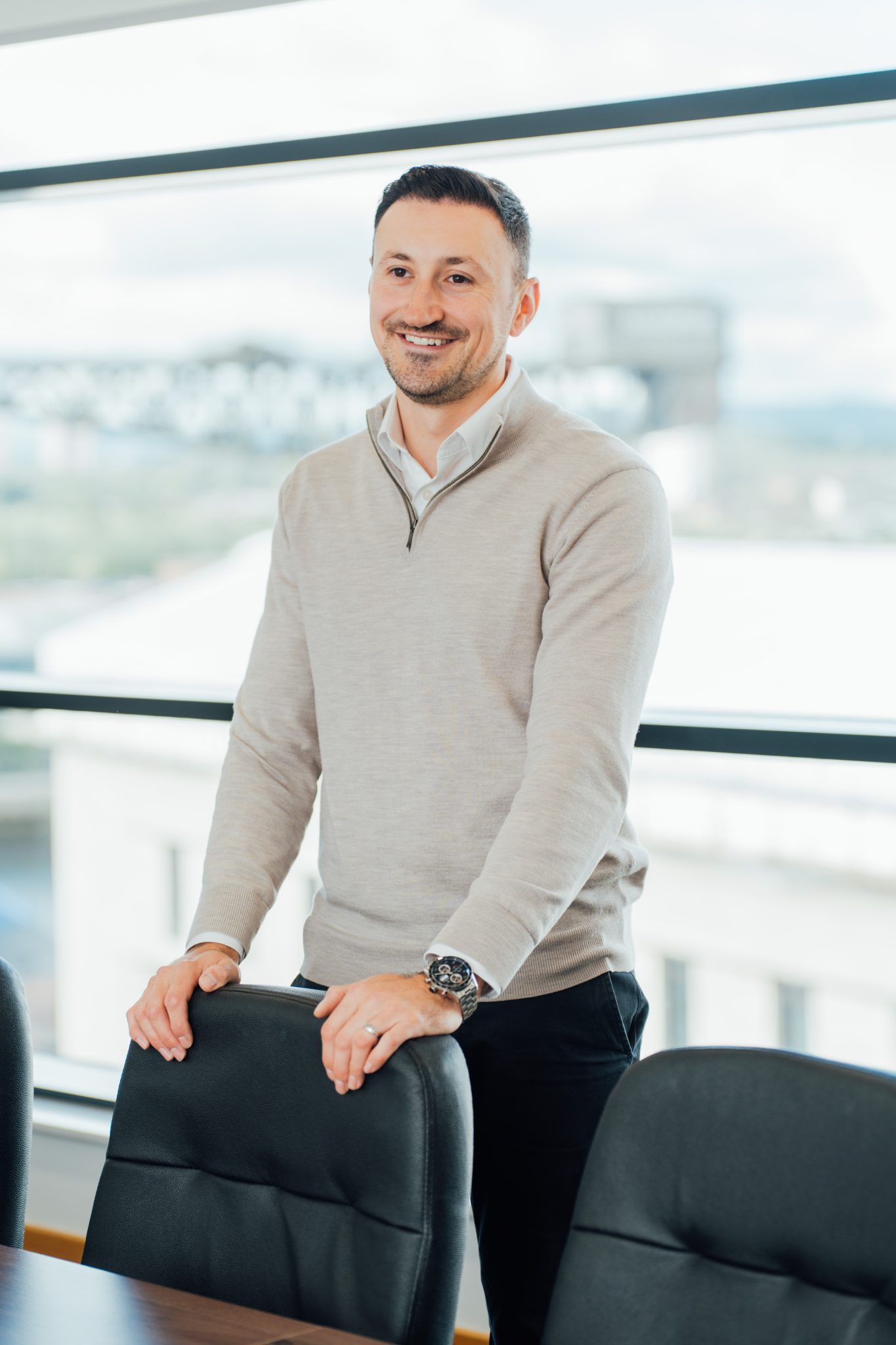 An image of Alistair Stewart wearing a grey jumper and standing in front of a chair smiling at someone.