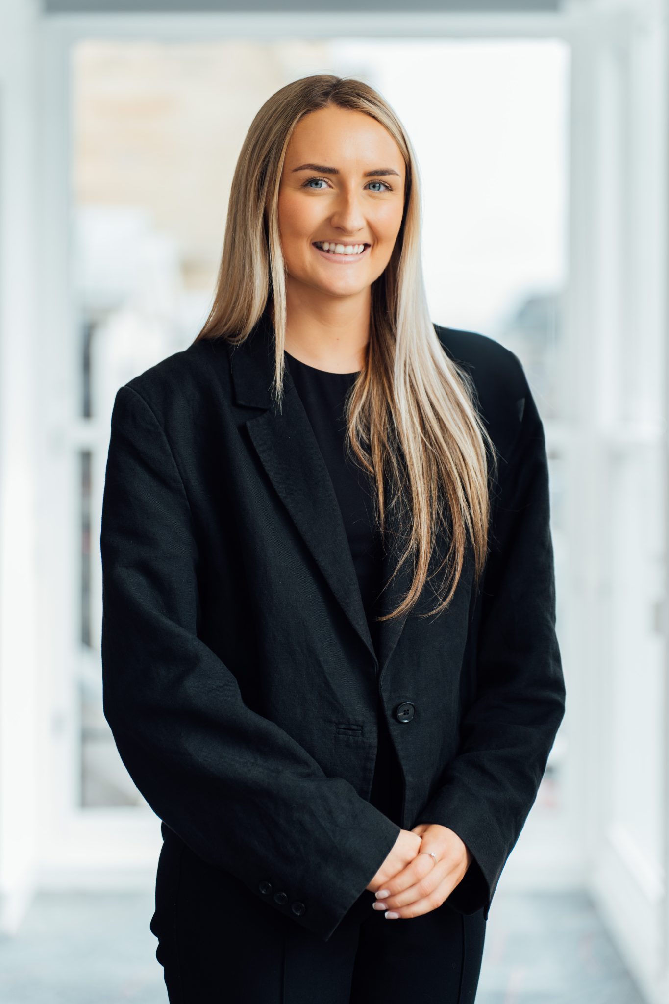 An image of Emily Ross wearing a black blazer and standing next to a wall smiling.