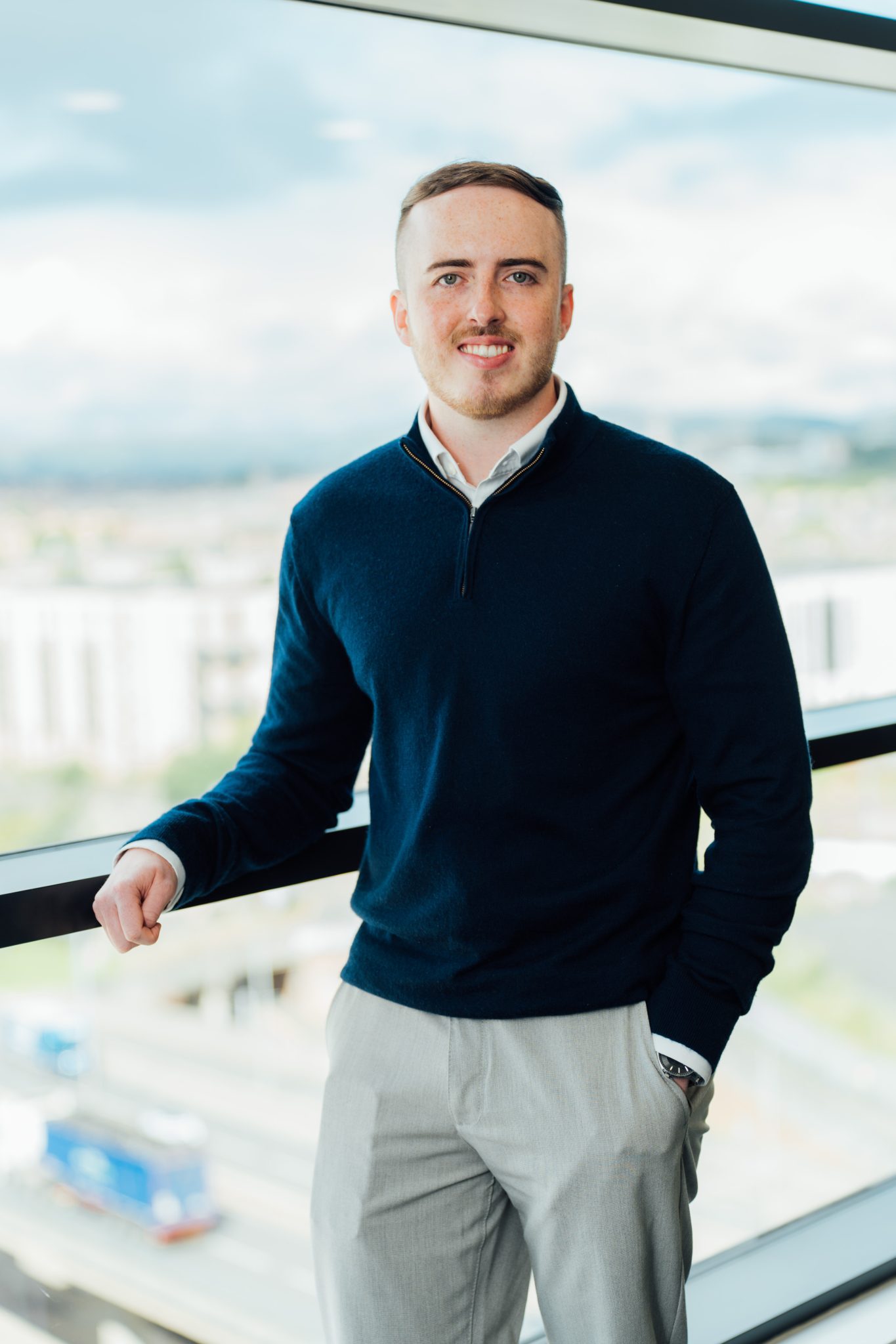 An image of Lewis Handley standing next to the window wearing a shirt jumper and cream trousers. His hand is in his pocket.