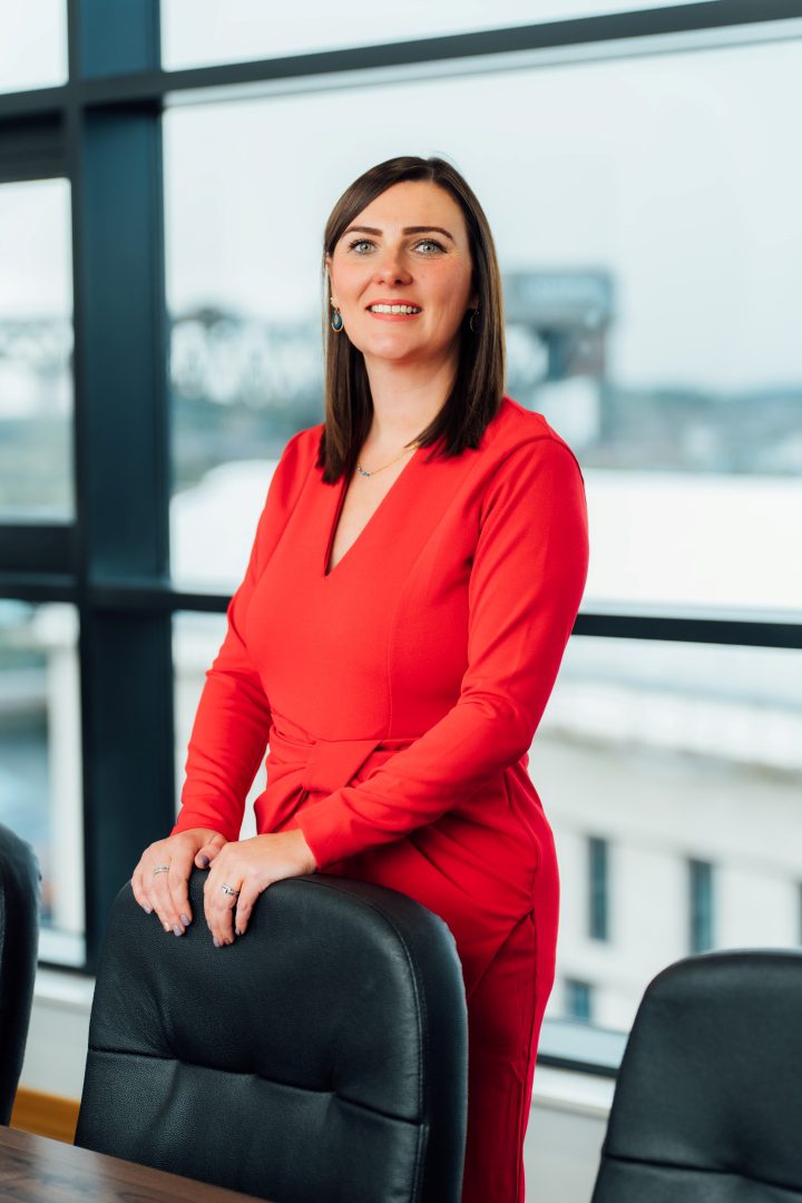 Natalie O'Hare standing behind an office chair by the window in the Glasgow office boardroom. she is smiling at the camera