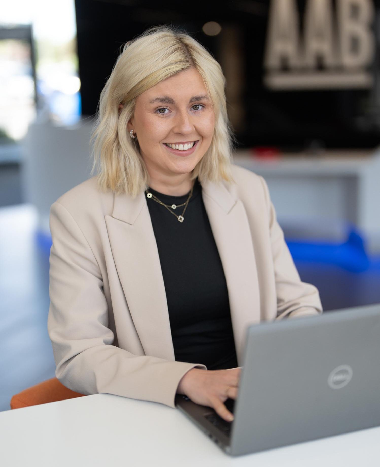 Chloe Singer sitting at a table in the office reception typing on her laptop smiling at the camera
