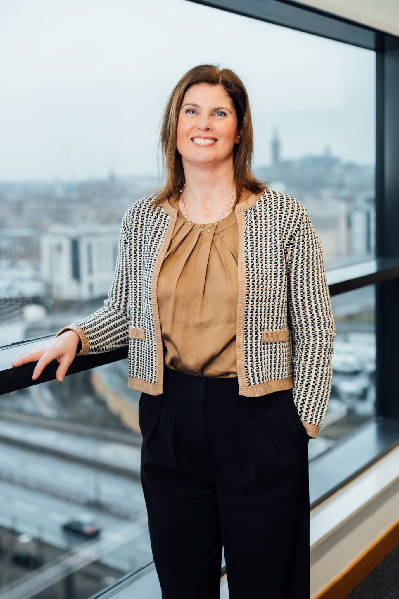 An image of Lorraine Quinn wearing a gold top with a cardigan. She is standing in the Glasgow office next to a window.