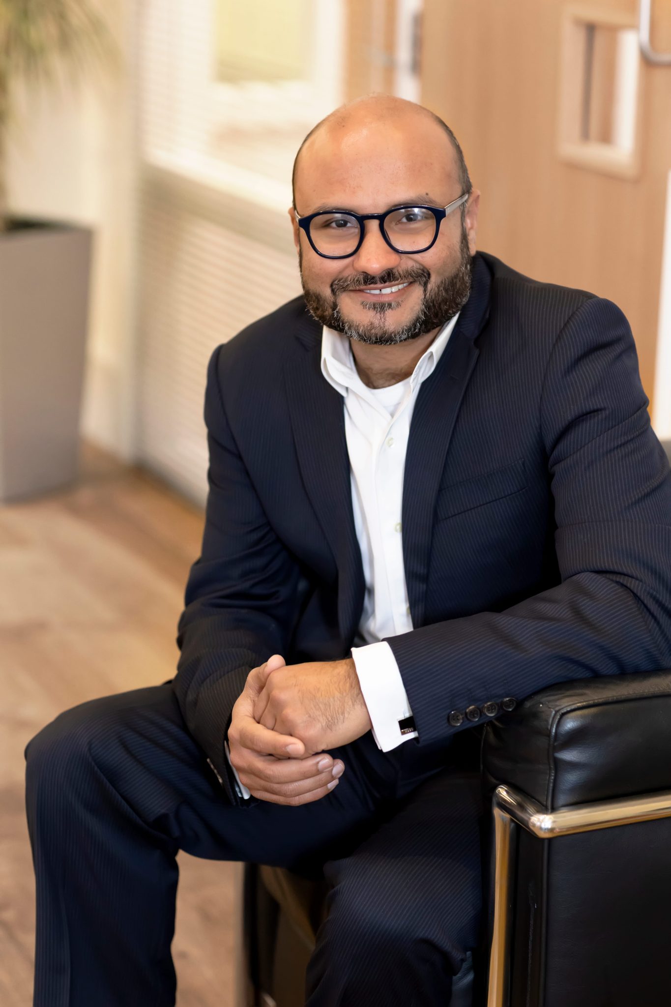 An image of Muhammad Sharif wearing a black suit with a white shirt. He is sitting on a chair smiling at the camera.