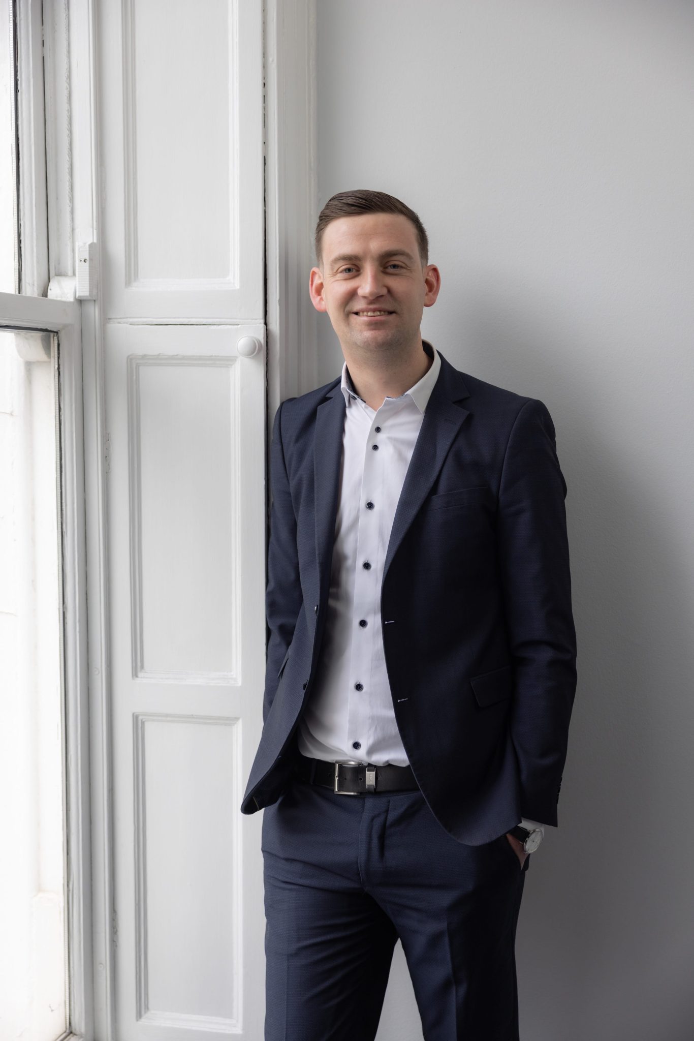An image of Brian Dunne wearing a suit and shirt. He is smiling at the camera and leaning against a wall.