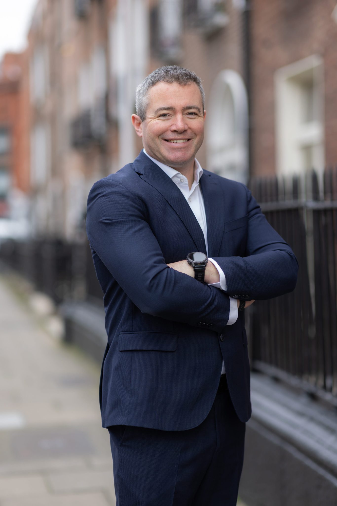 An image of Colm Duggan wearing a suit and tie. He is standing outside with his arms crossed.