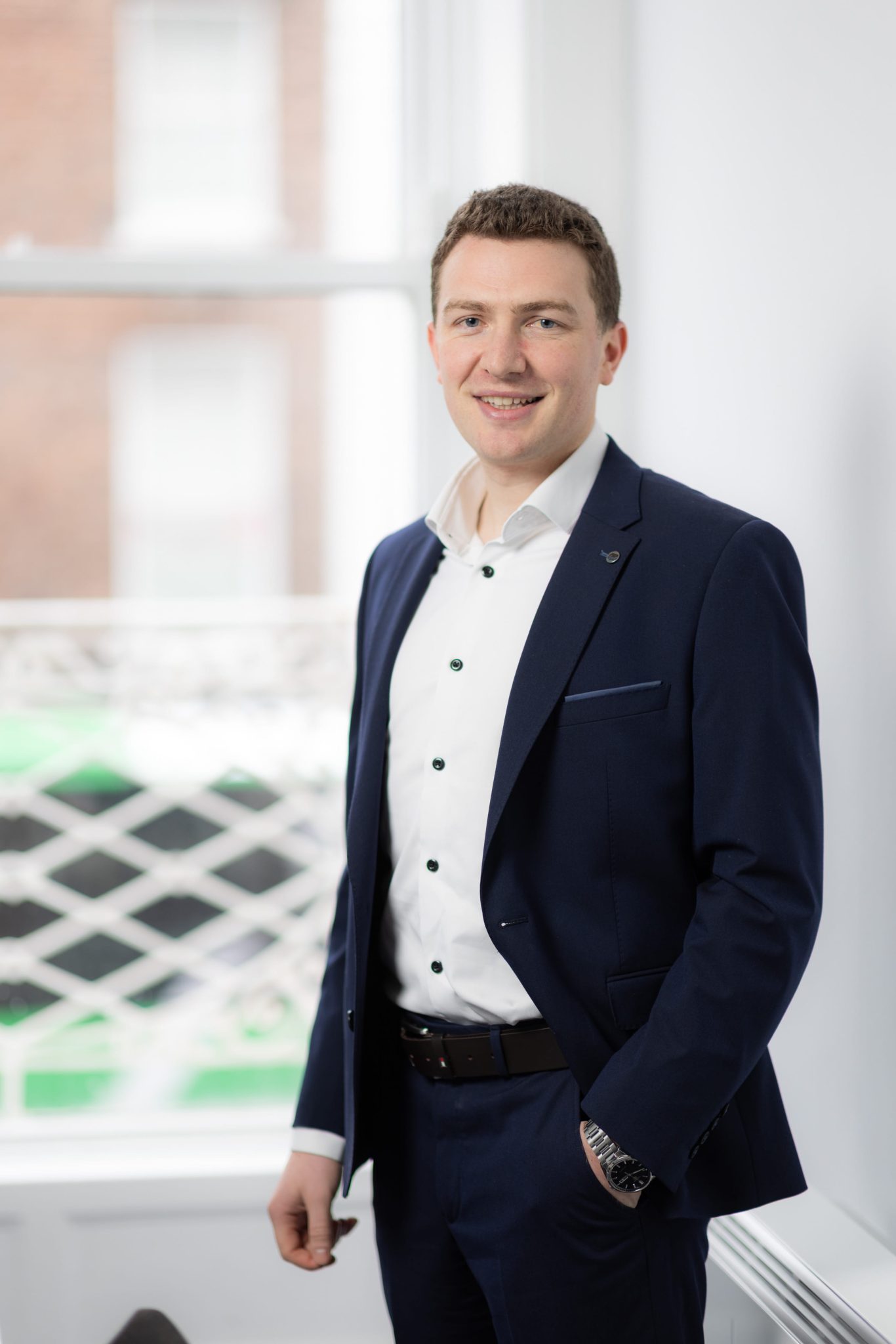 An image of Ruairi Power standing in the office wearing a black suit jacket and a white shirt. His hand is in his pocket.