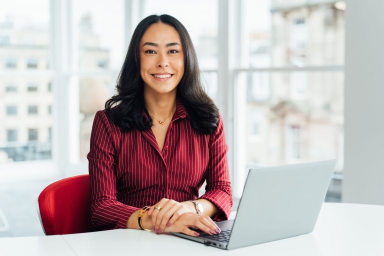 An image of Hannah Wildsmith wearing a burgandy top and smiling at the camera. She is sitting a desk with her laptop.