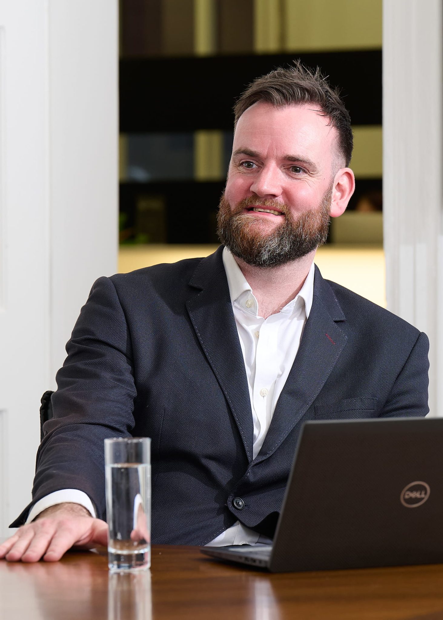 An image of Robert Wilson wearing a suit and shirt. He is sitting at a desk with his laptop.