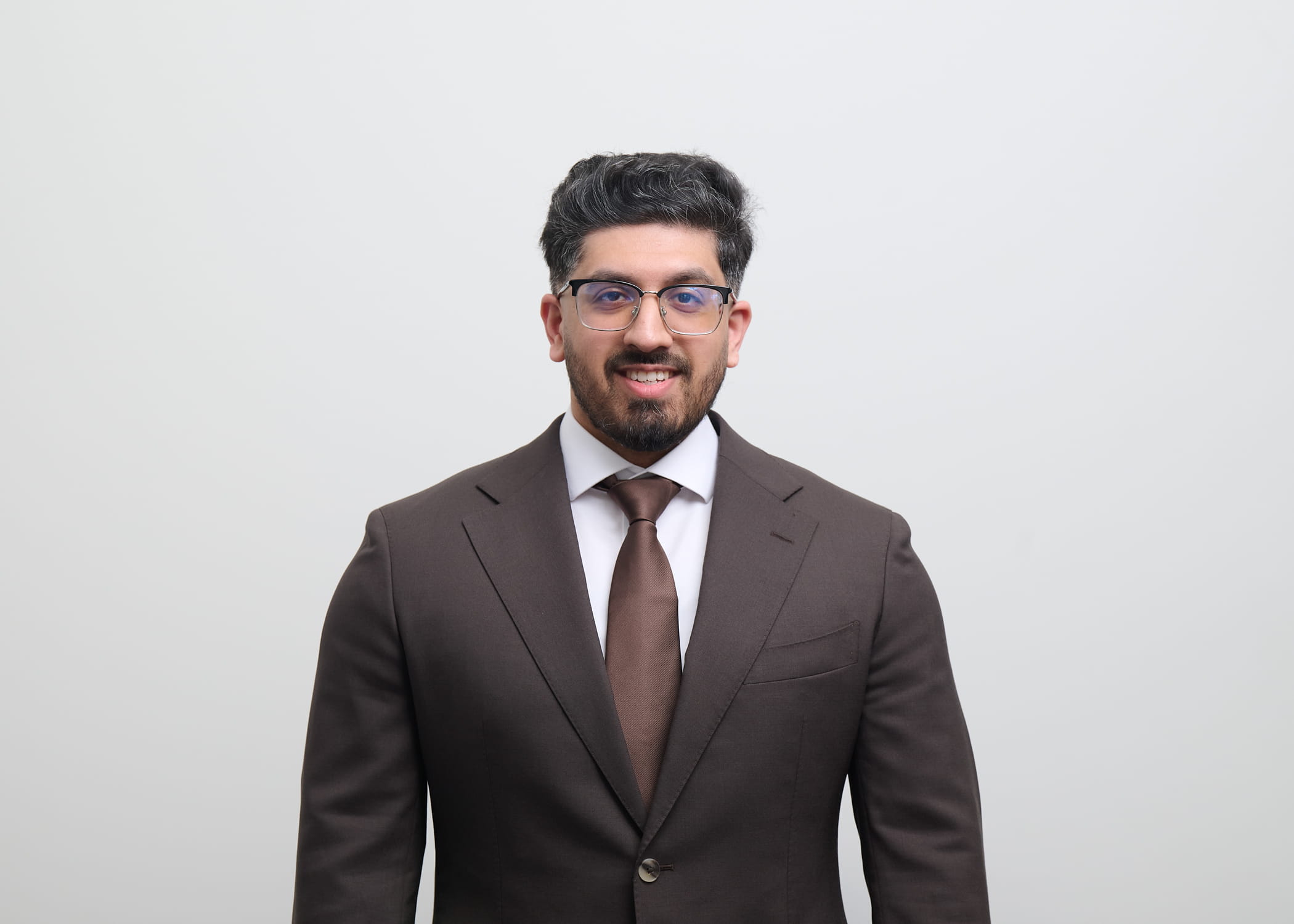 An image of rafiq Gazi smiling at the camera. he is wearing a brown suit and brown tie.