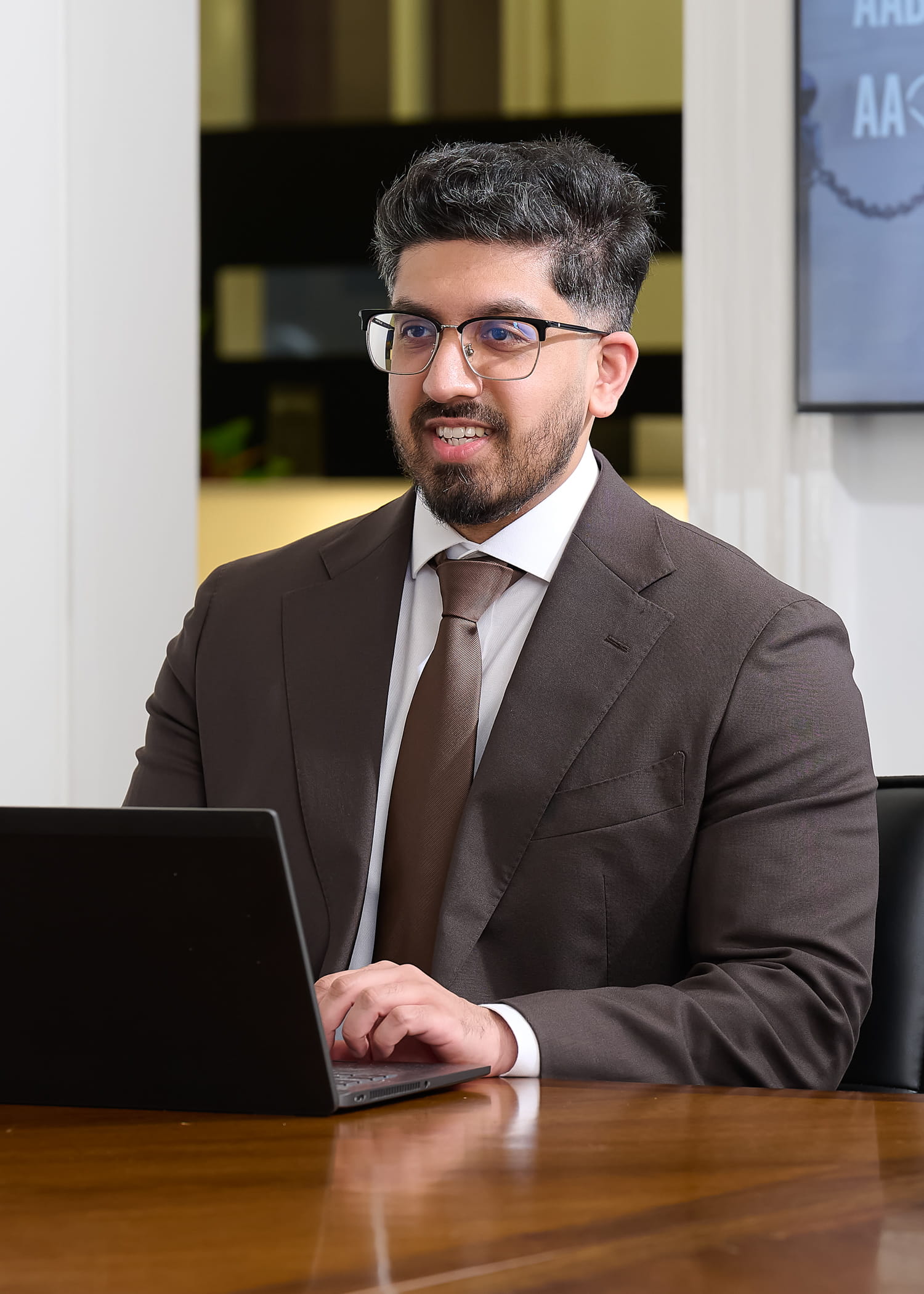 An image of Rafiq Gazi wearing a brown jacket and brown tie. He is sitting at a desk with his laptop.