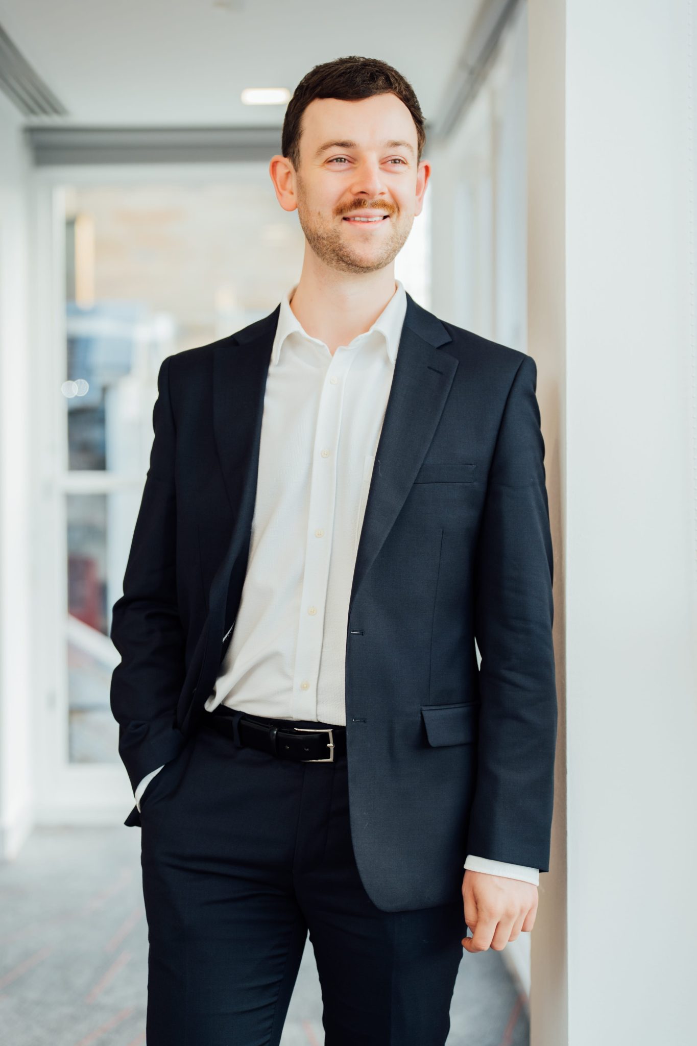 An image of Steven McCormick wearing a black blazer with a white shirt. He's leaning against a wall and looking out the window.