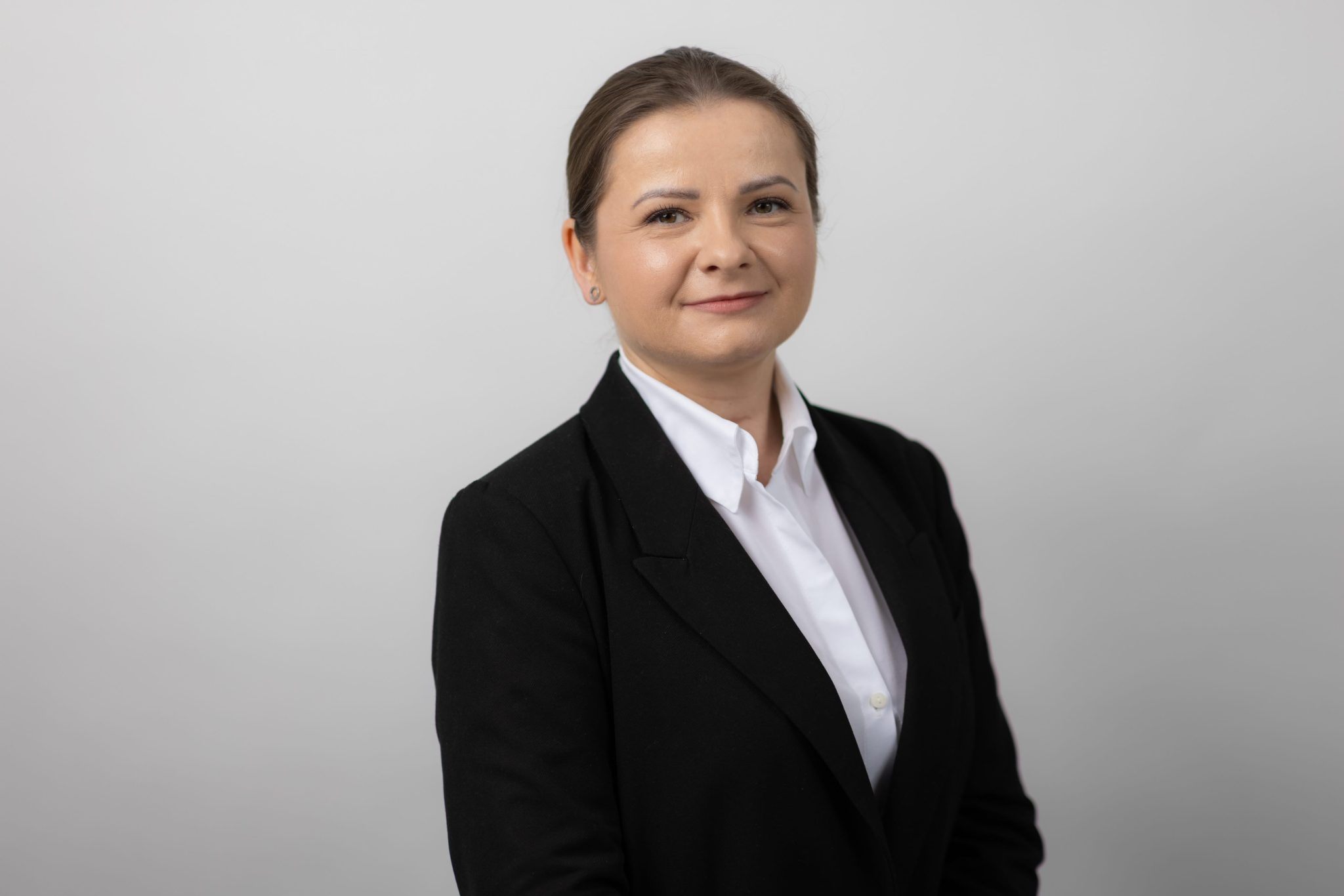 An image of Zoe Podraza Ostrega wearing a black suit jacket and a white shirt. She is smiling at the camera.
