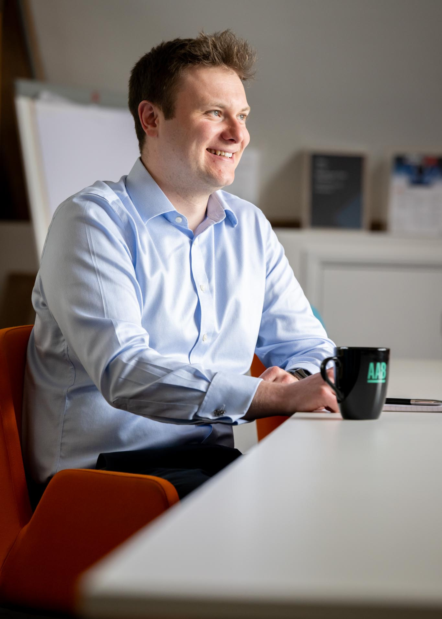 Cellan Richards of AAB sitting at a white table with an AAB mug, wearing a blue ling sleeve shirt, black trousers and sitting next to red chairs