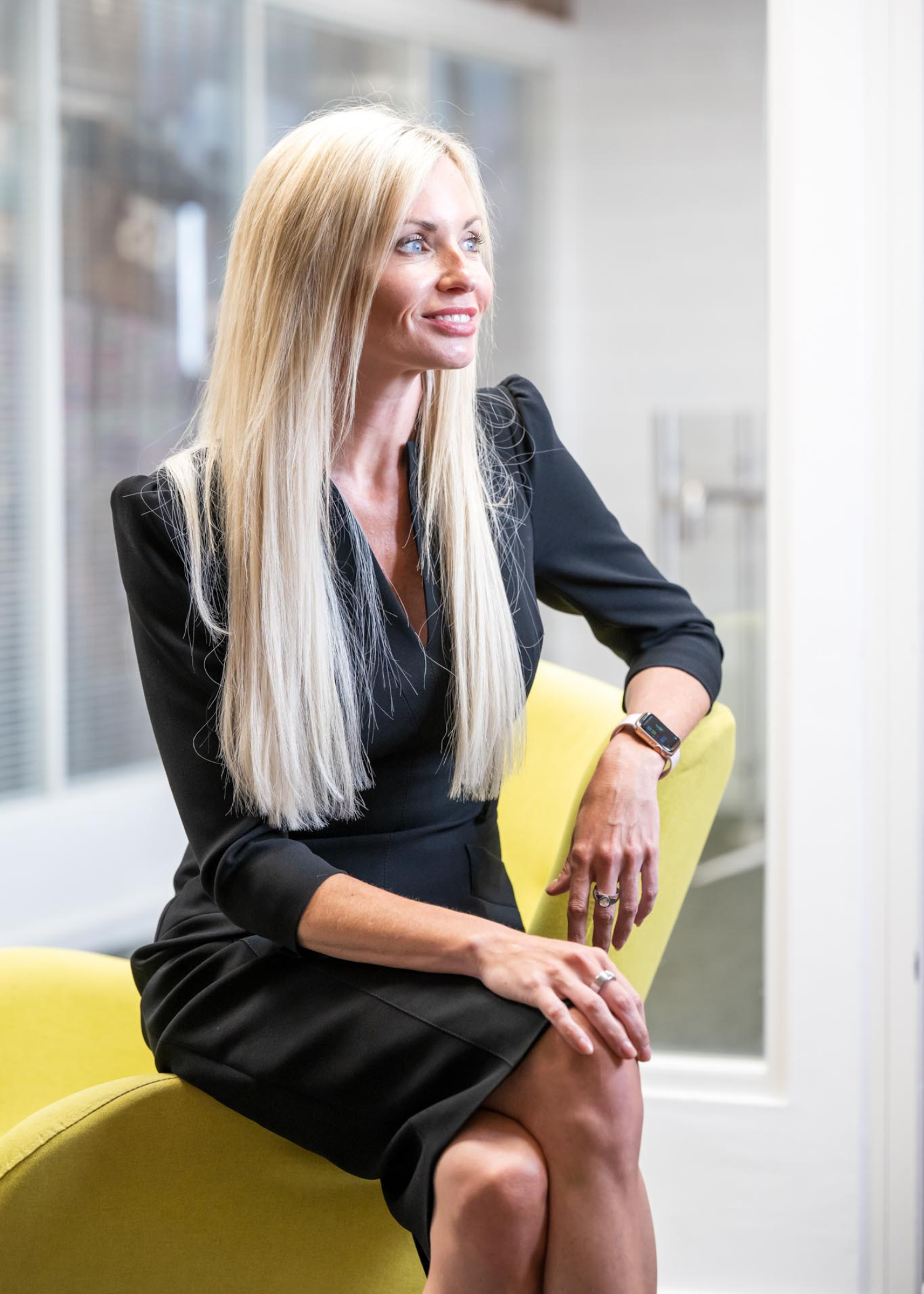Leanne Thomas, Corporate Director at AAB, has long blonde hair and is wearing long sleeved black dress. The dress has a slight raised/padded effect on the shoulder. Leanne is sitting on the arm of a yellow chair with a glass window behind her.