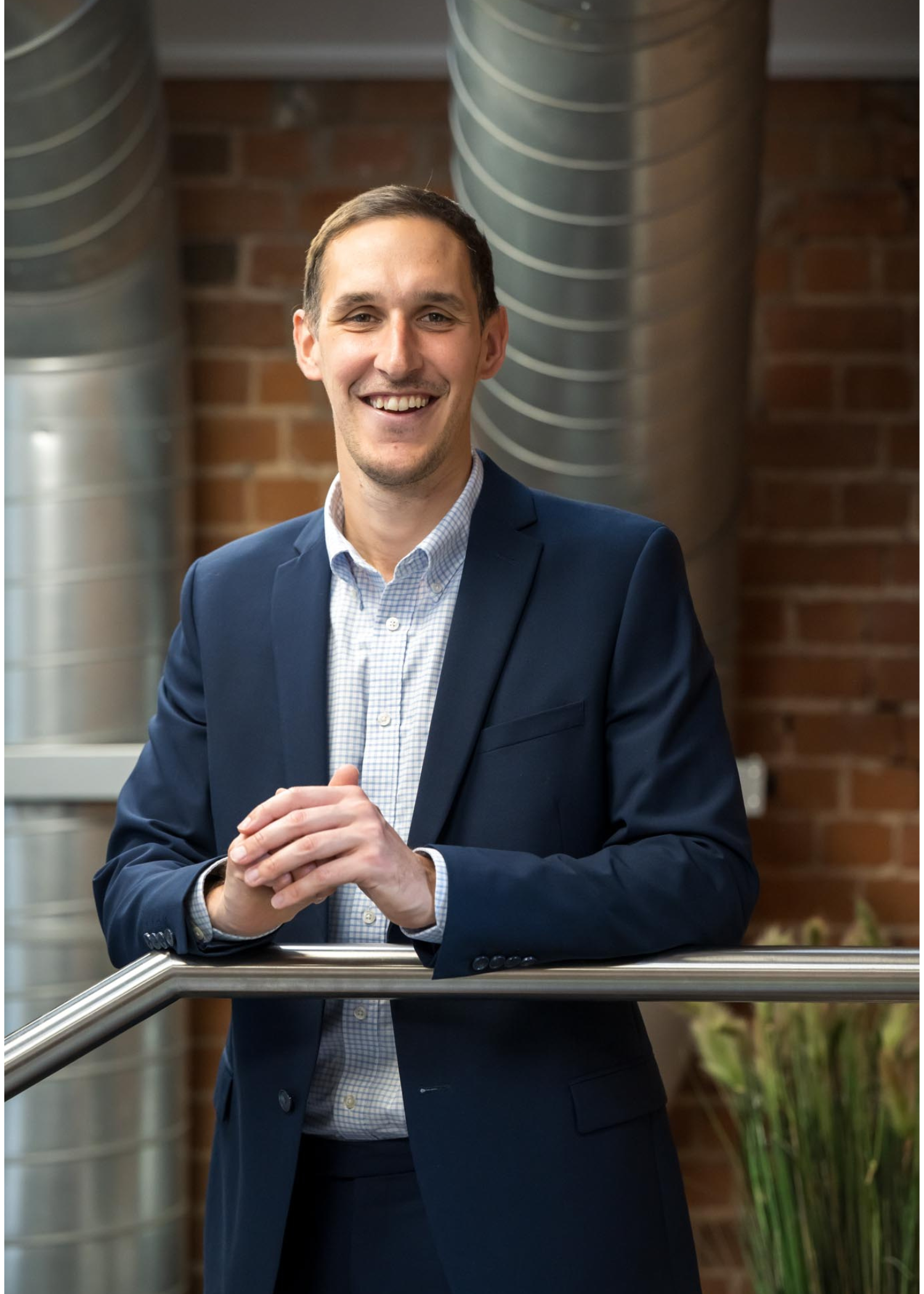 Joseph Sharp, Corporate Finance Director at AAB, is turned slightly to the left in his headshot photo. He has dark short hair and is wearing a dark blazer with dark trousers and a white shirt. He is posed facing the camera and standing on the top of the metallic staircase with his arms leaning against the bar.