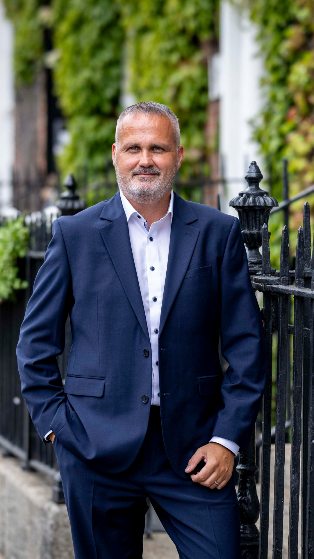 Des Barnes - Practice Director, Dublin AAB. Stood outside against black railings with foliage on the building walls.